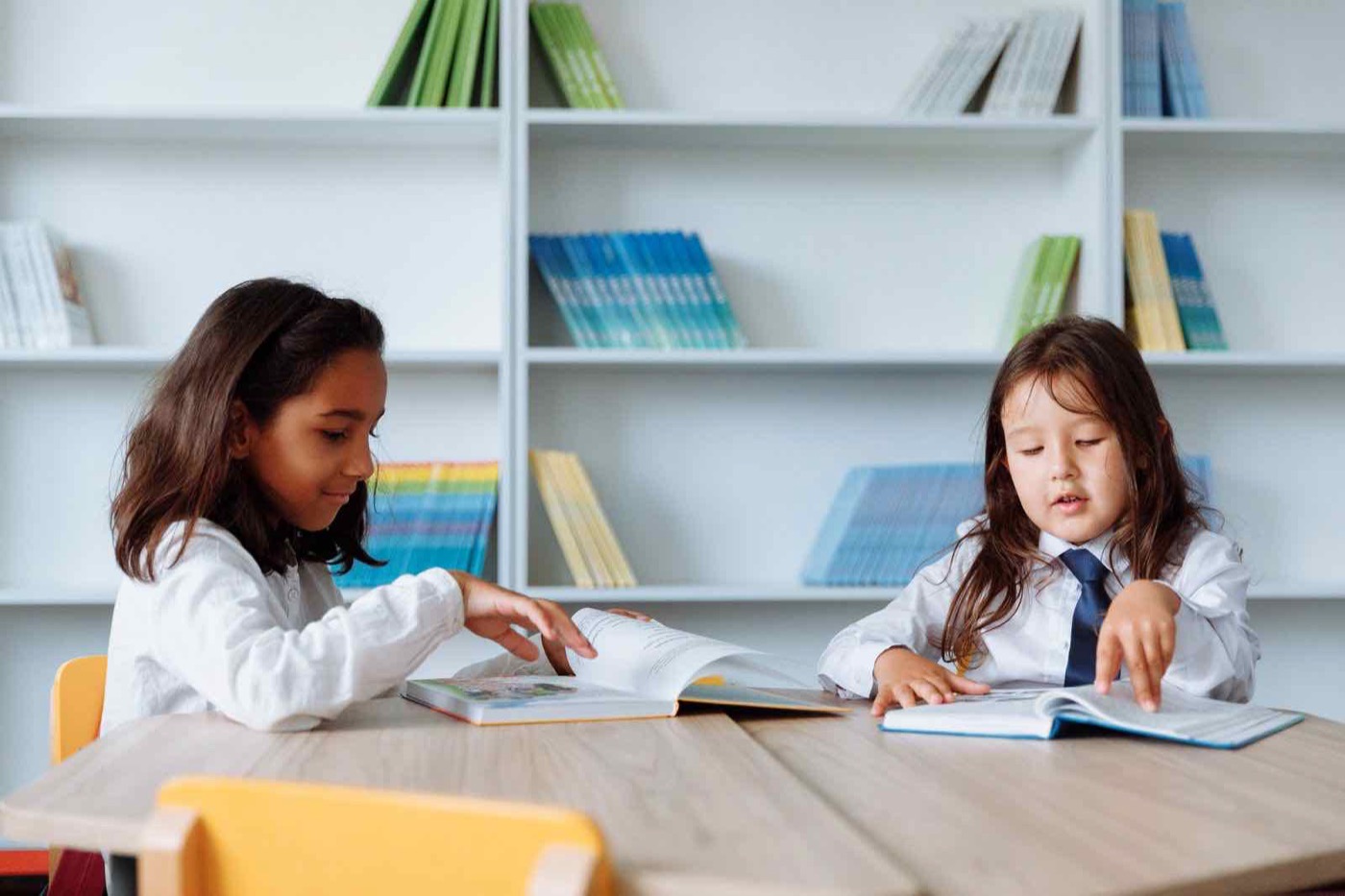 Two students at a table in a school library, one pointing something out in an open book to the other.