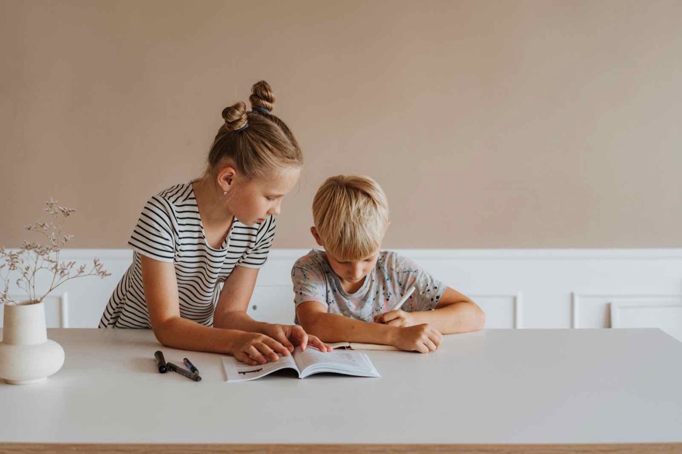 An older sister pointing out a step in a workbook while her younger brother writes the answer.